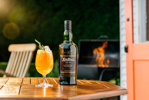 Bottle of Ardbeg An Oa whisky on a wooden table with a cocktail in the foreground, outdoor BBQ setting.